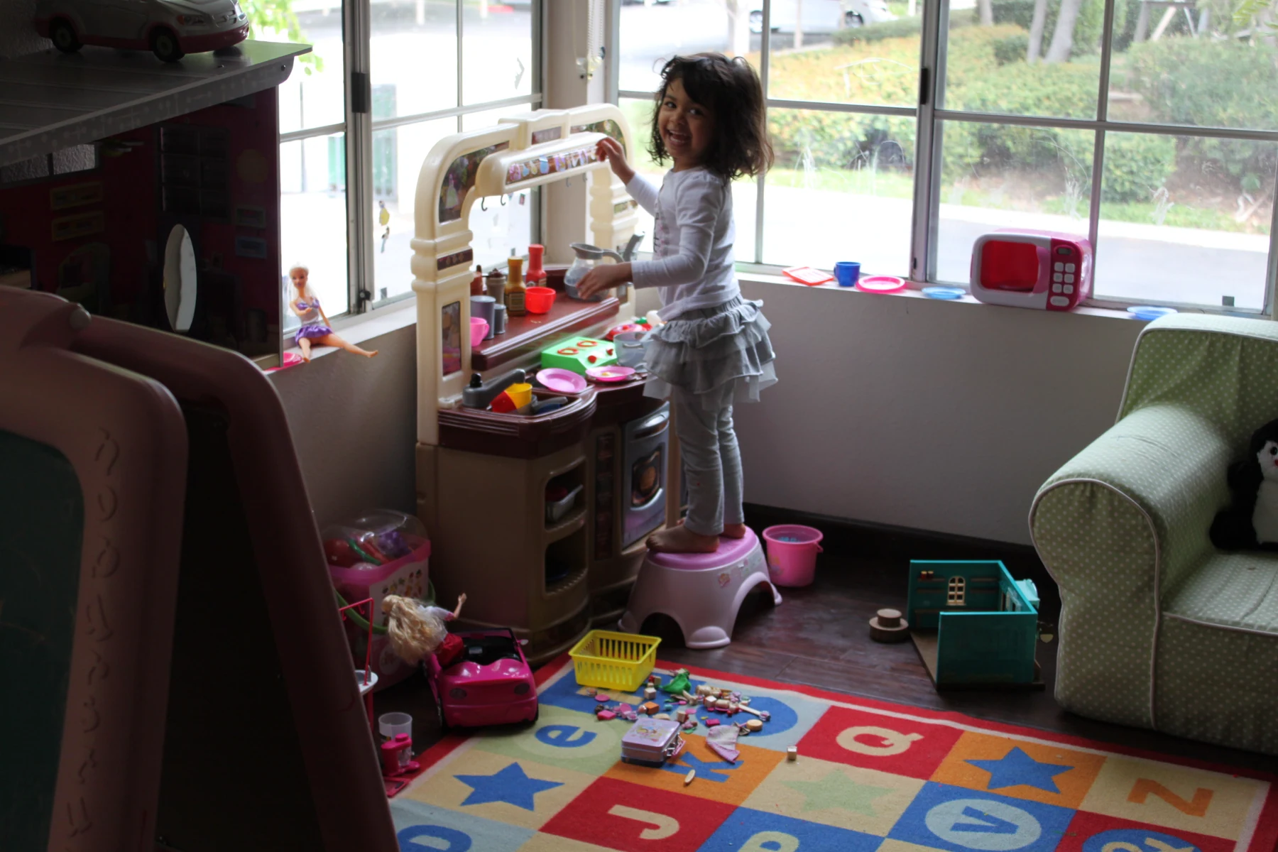 Mili, age three, stands on a pink step stool at her toy kitchen.