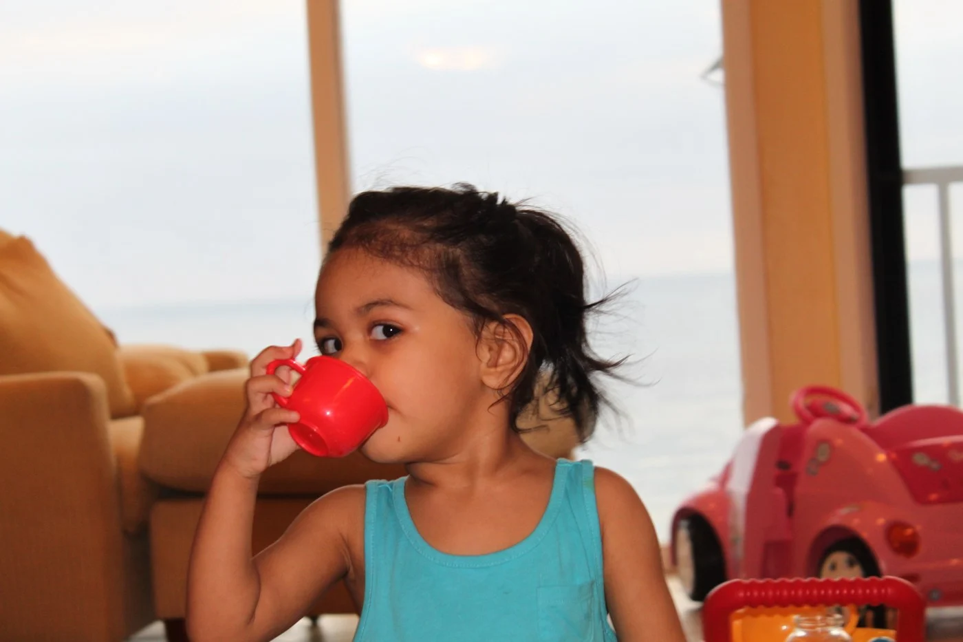 A child drinking from a small red cup near a toy kitchen.