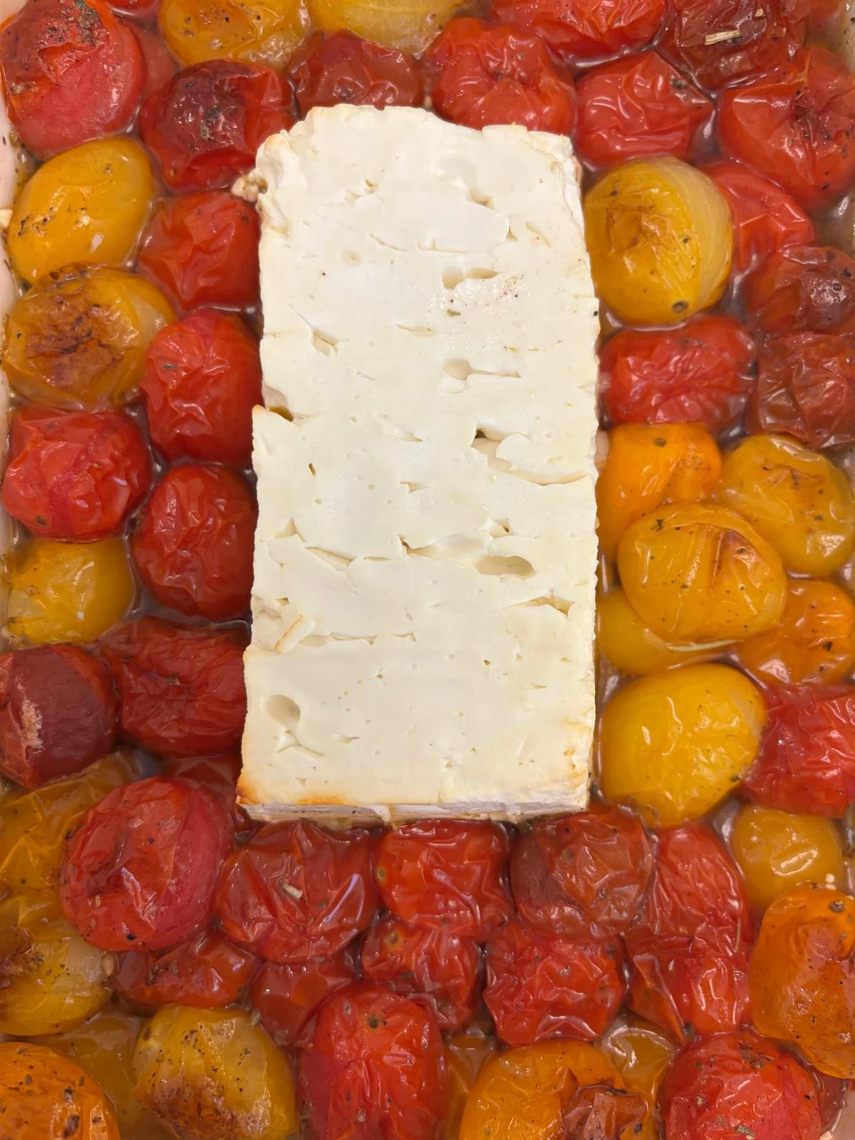 Rectangular white cheese block in roasted red and yellow cherry tomatoes with oil.
