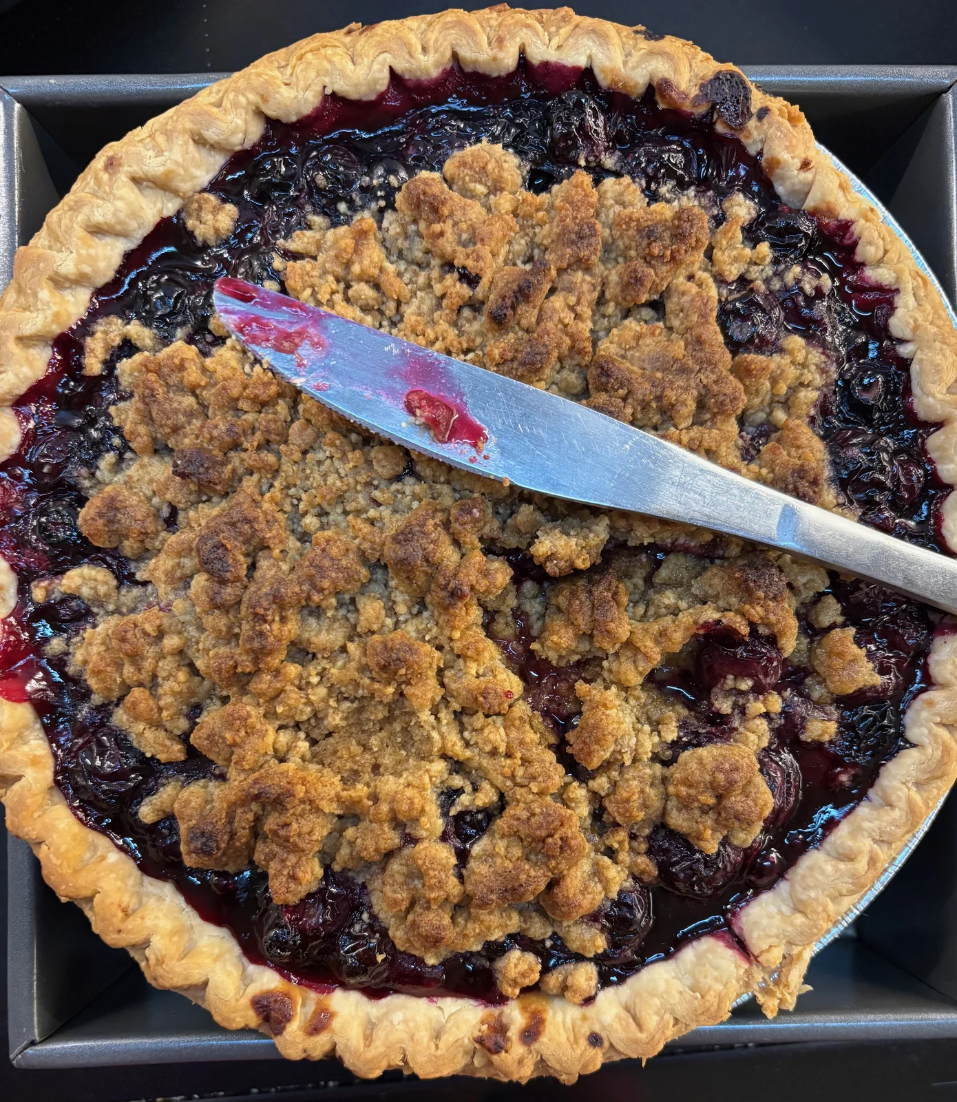 Crimped single pastry crust, dark red-purple whole/halved fruit and golden streusel topping. Large round wrinkled skins look more like cherries than blueberries.