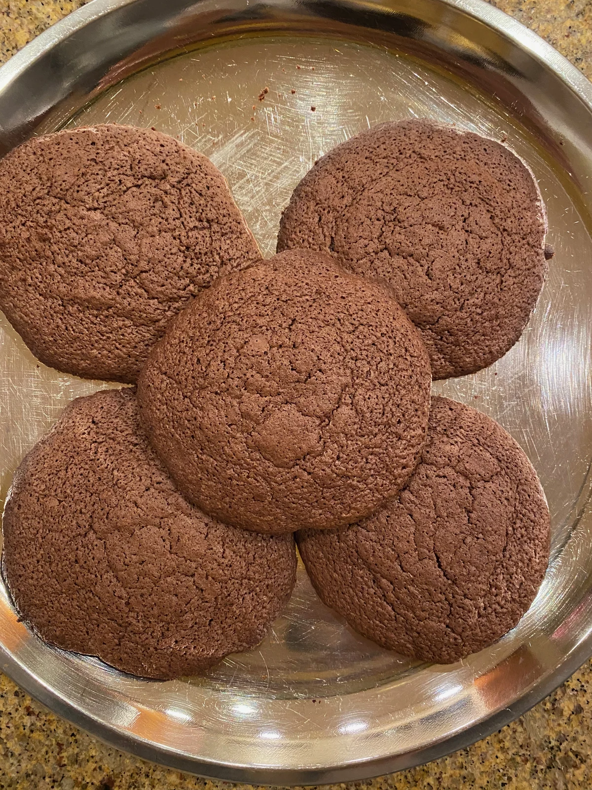 Five broad brown cocoa-colored cookies with fine porous tops and small cracks, on a steel plate.