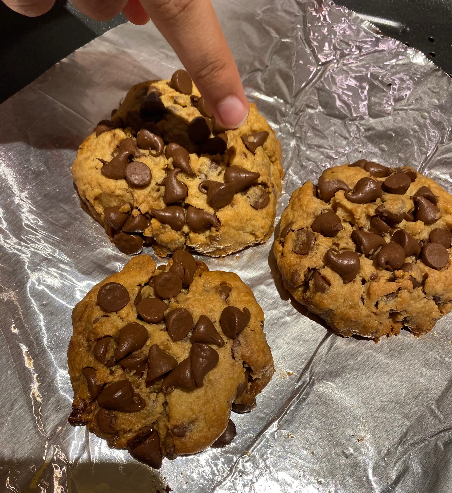 Three thick cookies with abundant surface chips and a pointing finger, matching existing loaded-cookie composition.