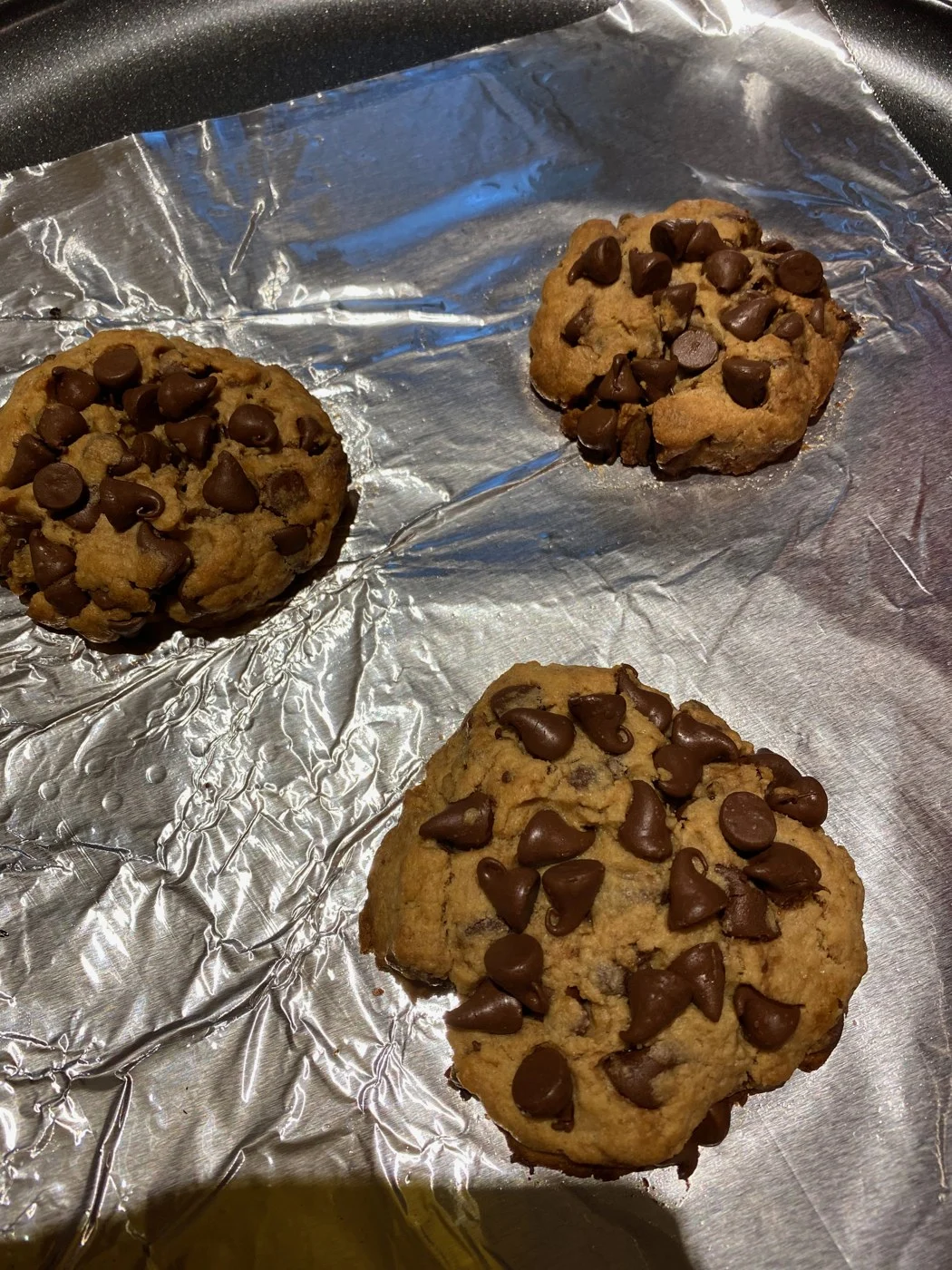 Three large chocolate-chip cookies spaced on a foil-lined baking tray.