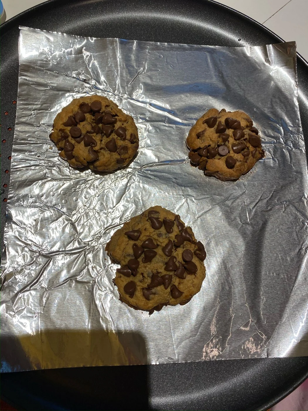 Three golden chocolate-chip cookies cooling on foil beneath a kitchen light.