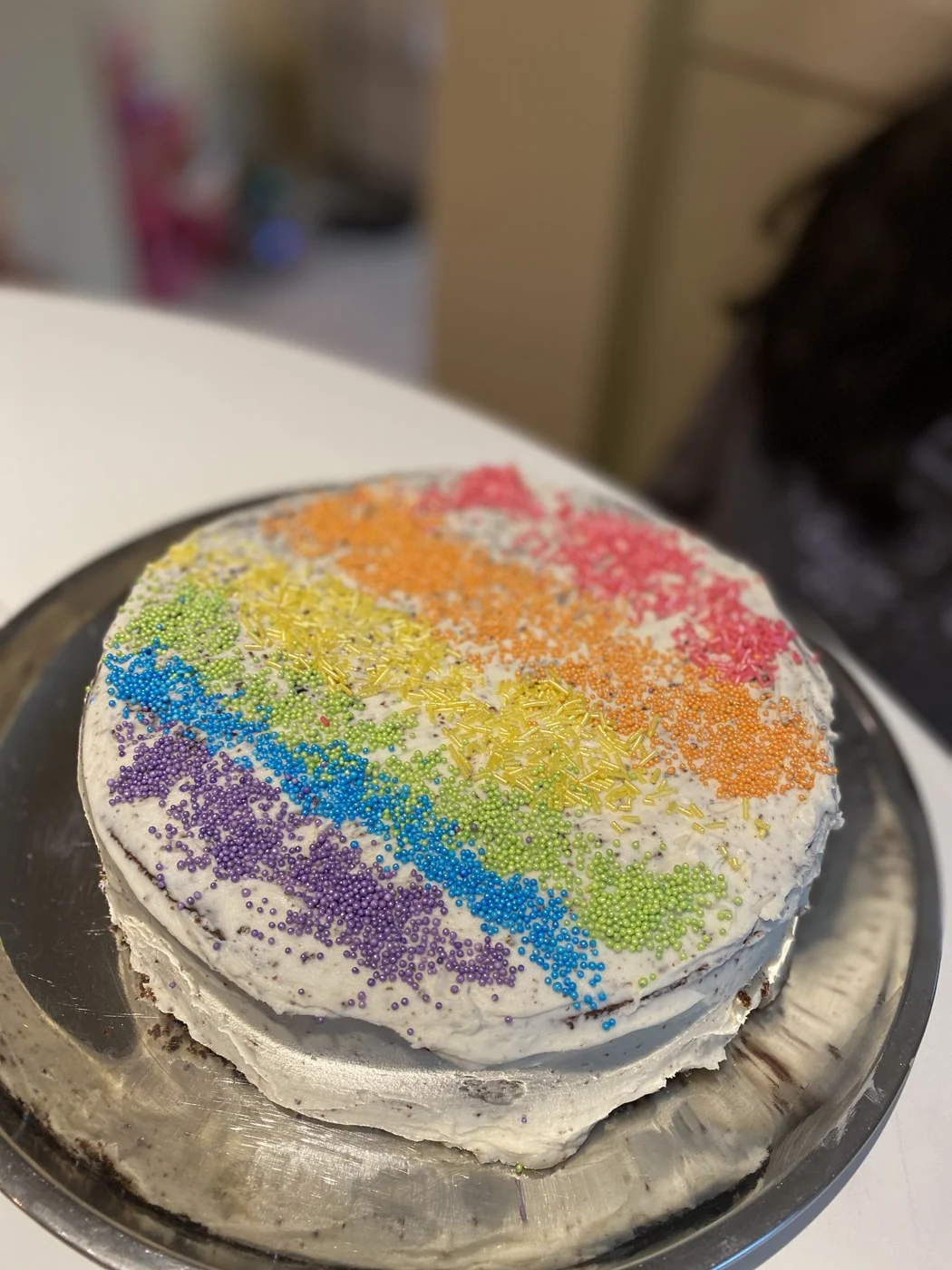 A round frosted cake covered with rainbow stripes of sprinkles.