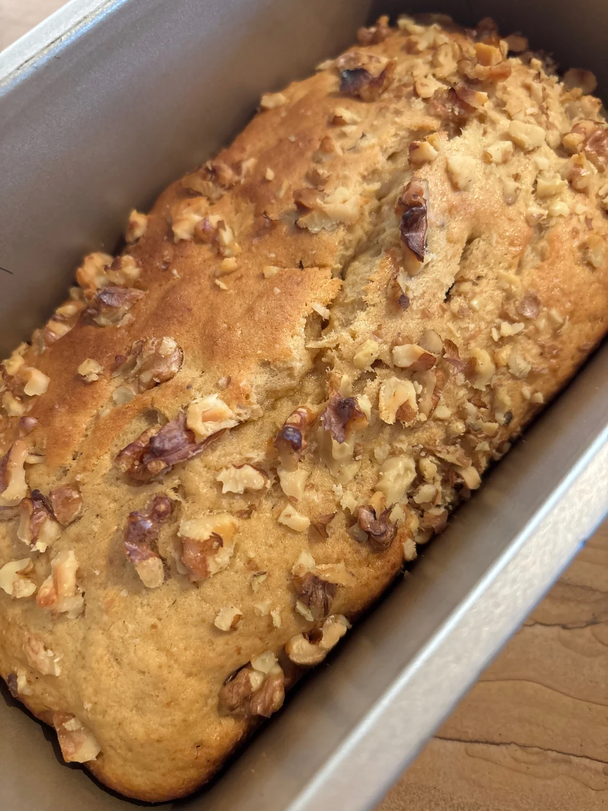 Golden cracked loaf in rectangular tin with chopped walnuts on top and nut flecks in the crumb.
